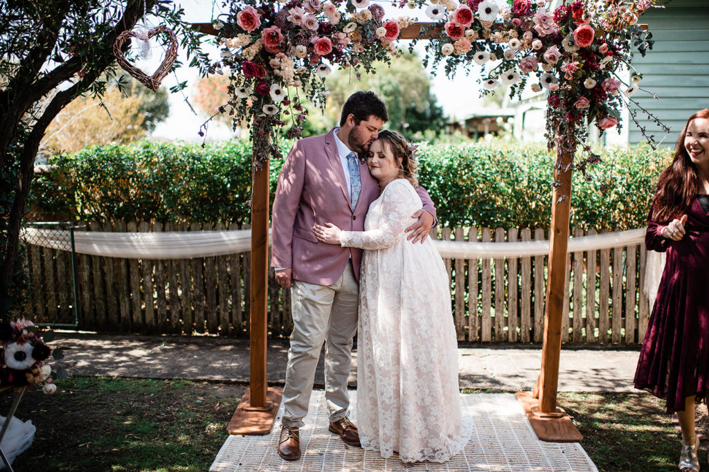 couple embrace in front of arbour decorated with florals at New Lambton wedding