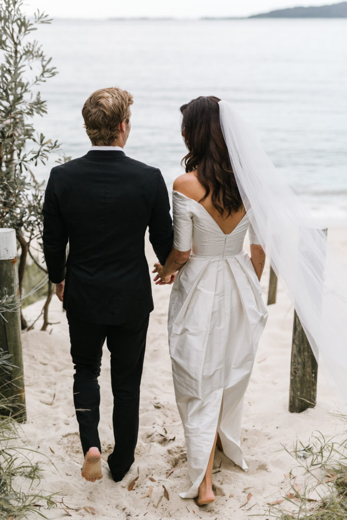 Couple in black tie and barefoot walking on the sand for their Shoal Bay Beach Elopement 