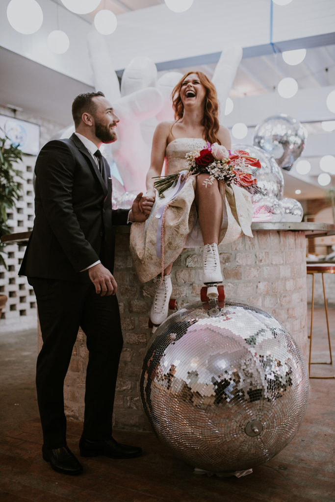 Bride sitting on the bar with her rollerskates on leaning on a huge disco ball talking to groom who is leaning on the bar