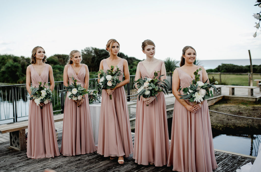 Bridesmaids in pale pink dresses holding greenery bouquets at Caves Coastal Bar & Bungalows
