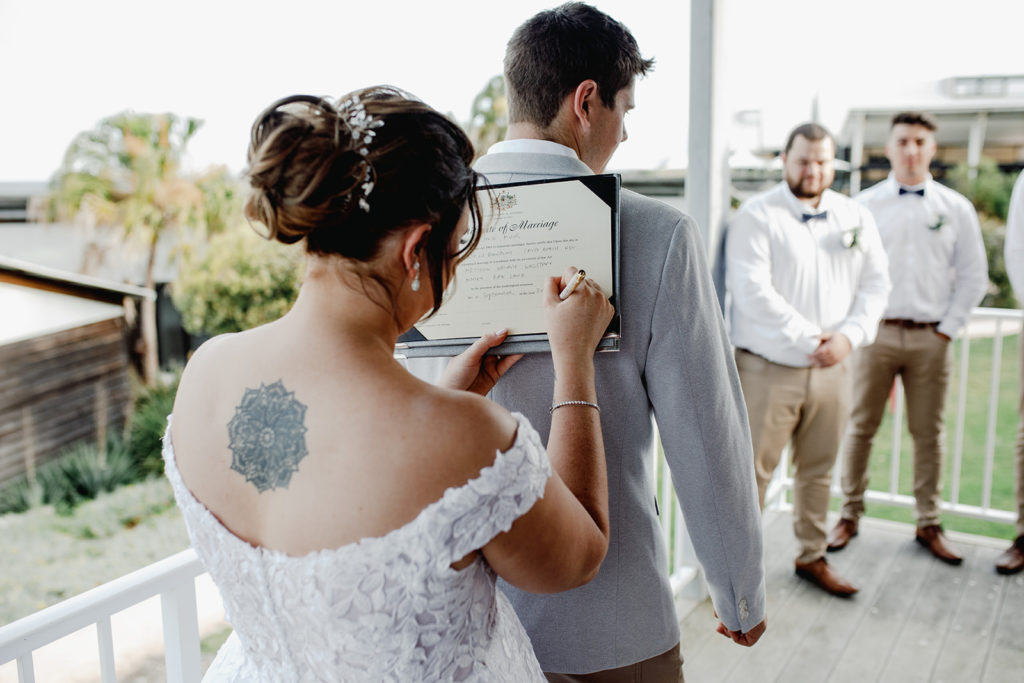 Bride with large back tattoo signs marriage paperwork while leaning on her husband's back at Caves Coastal Bar & Bungalows
