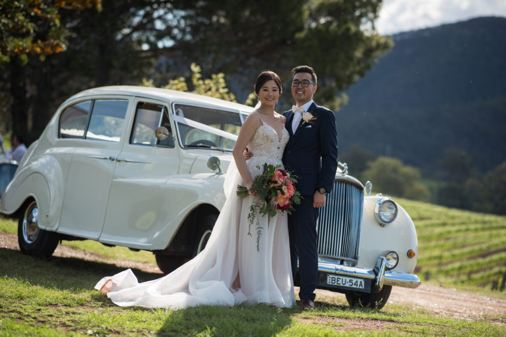 newlyweds smiling and posing in front of a white vintage car at a Hunter Valley Wedding Venue in Ben Ean