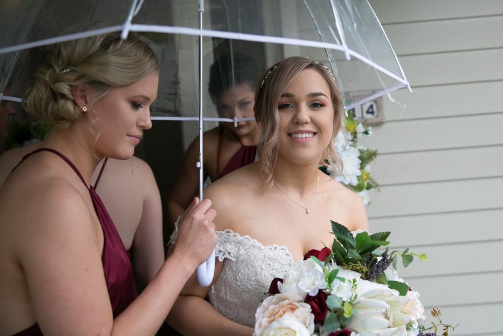 bride smiling at the camera beside bridesmaid holding up an umbrella