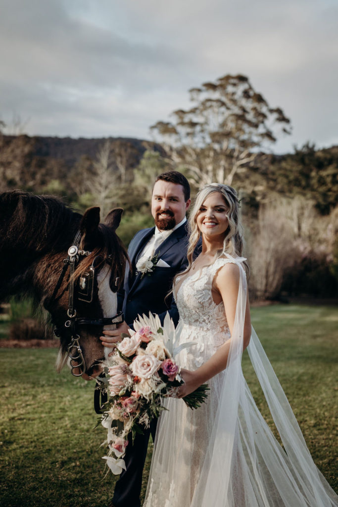 Bride and groom posing and looking at the camera beside a dark horse at Fernbank Farm