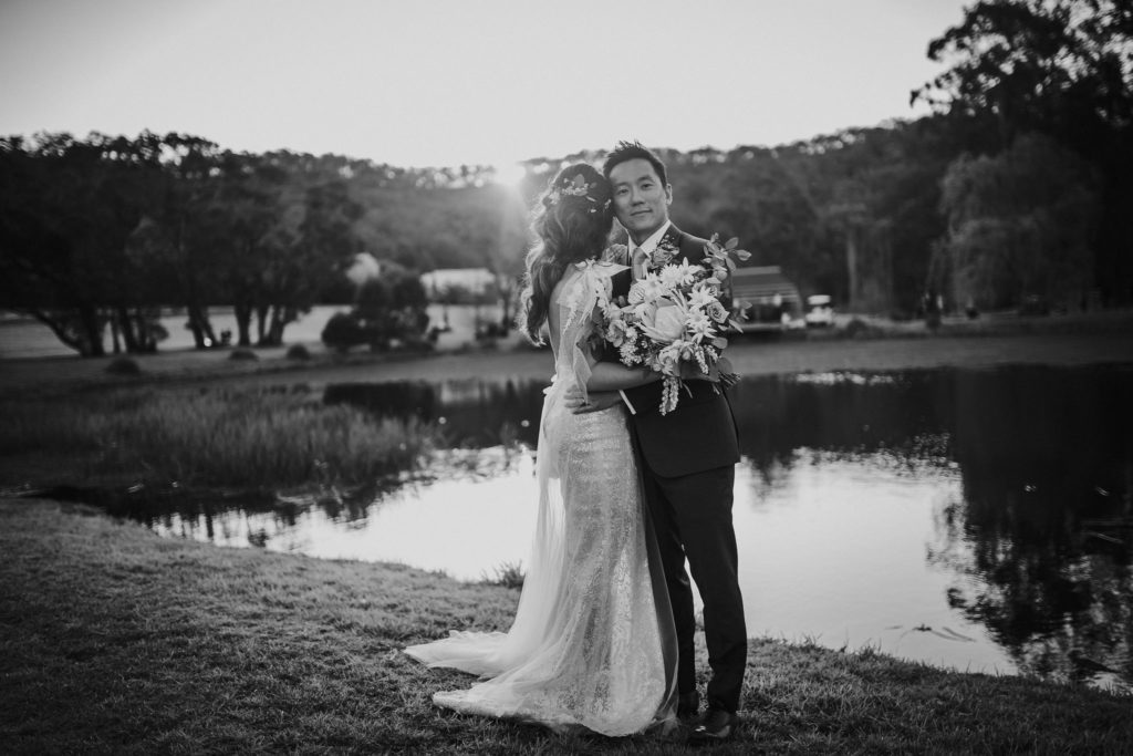 Black and white photo of the couple standing at the lake at The Woodhouse Wollombi. Groom looks towards the caera. Bride loosk away. 