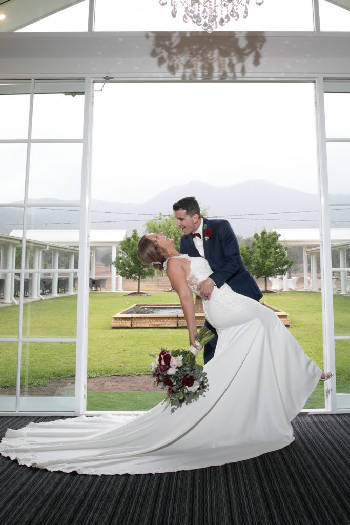 Groom romantically dip kissing bride with long dress train in front of the open French doors at Leogate Estate