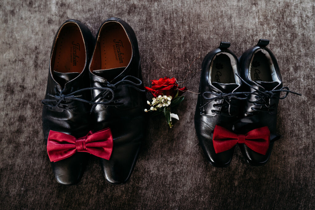 Groom and ring bearer's black shoes with red bow ties on them and a rose boutonniere between the two pairs