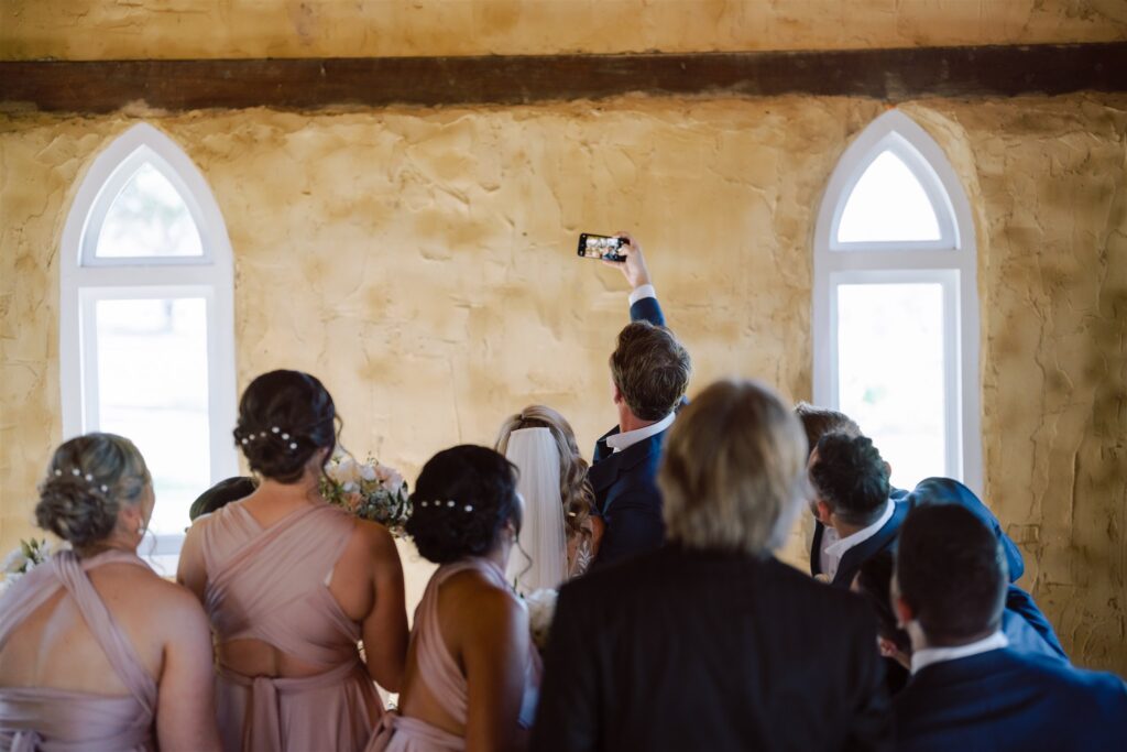 Groom raising his phone for a group with the rest of the wedding party, and guests at Peppers Creek Chapel & Barrell Room.