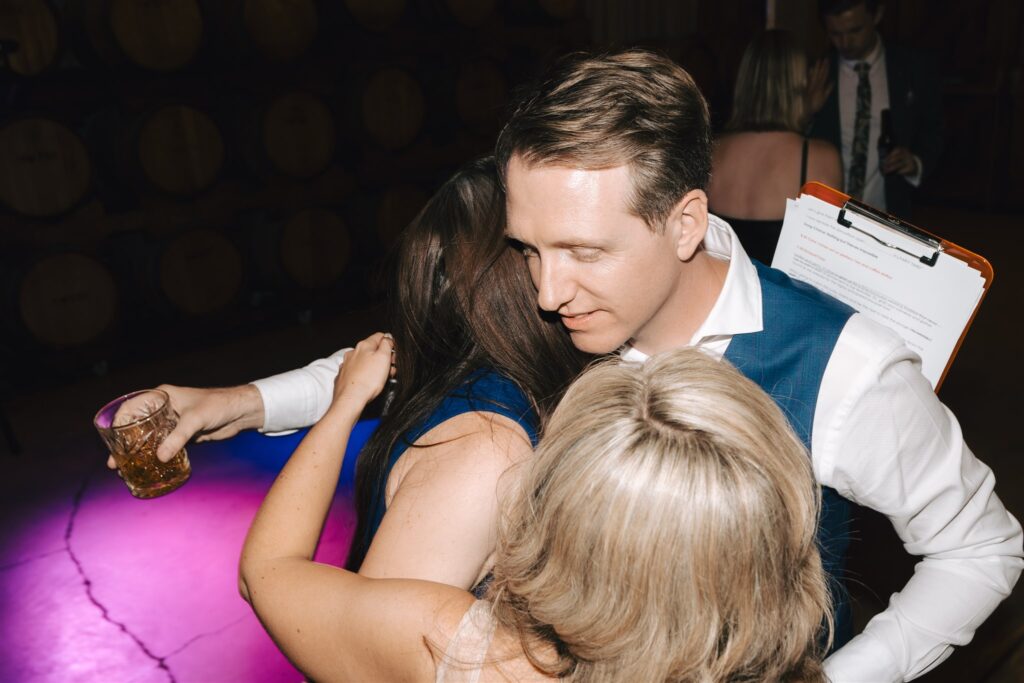 Groom hugging two wedding guests while holding a glass of alcohol