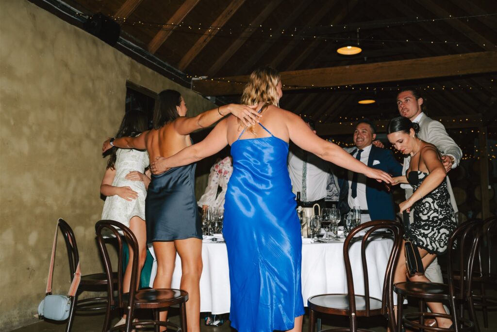 Some wedding guests forming a circle at a table with their arms on their waists, partying at Peppers Creek Chapel & Barrell Room.