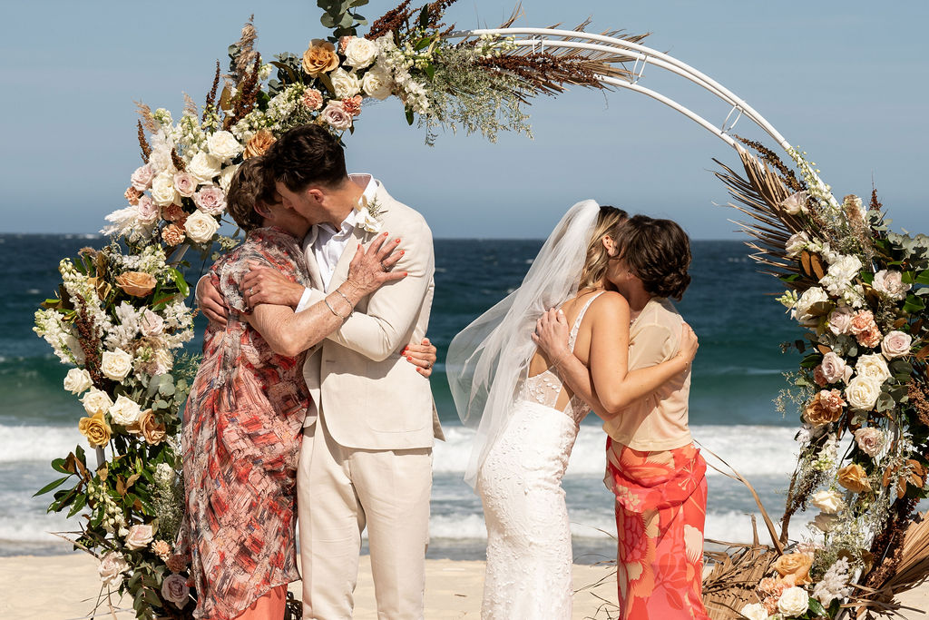Bride and groom each hugging their mothers in front of arbour at Zenith Beach