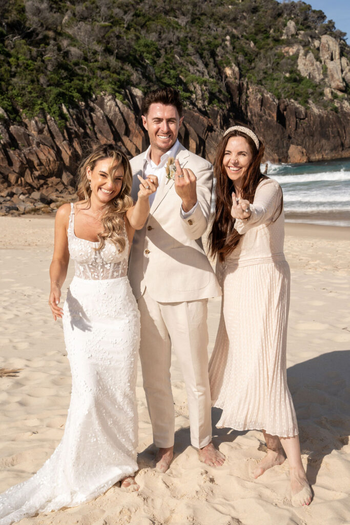 Celebrant Julie Muir and newlyweds smiling and showing their left hands' ring fingers at Zenith Beach