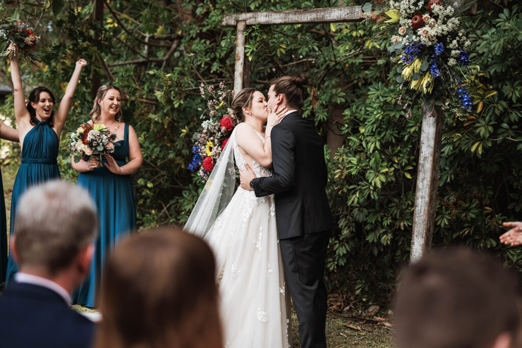 Bride and groom have first kiss at at Leaves and Fishes garden in front of an arch. 