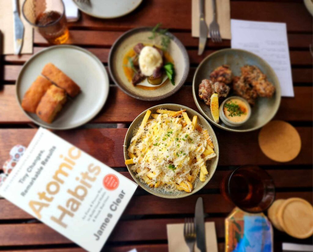 Atomic Habits by James Clear book on a table surrounded by lots of food including chips