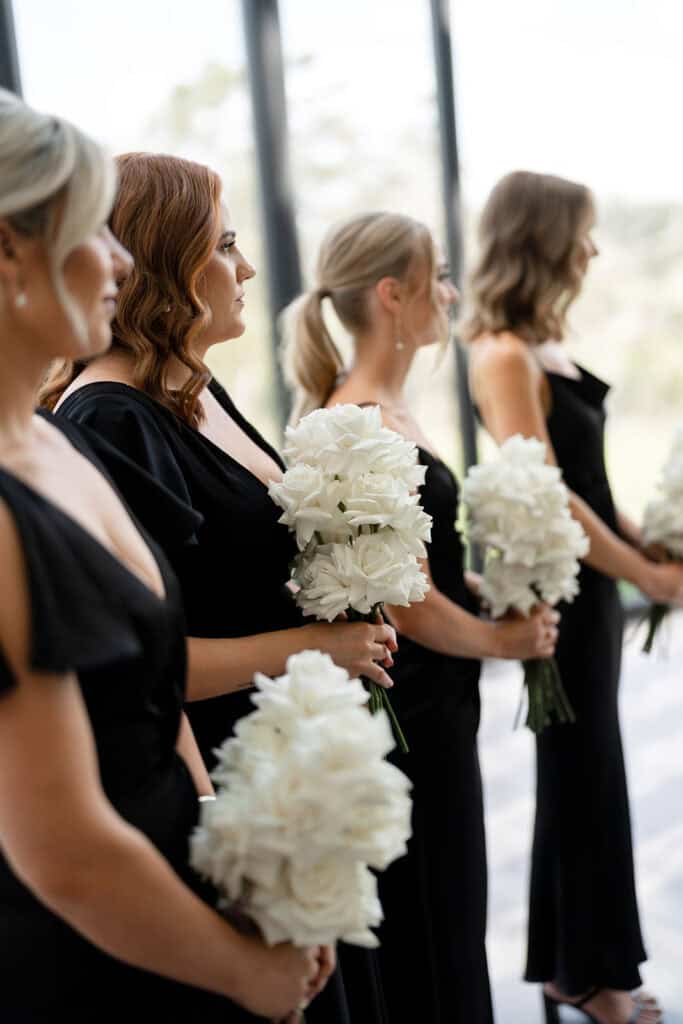 Bridesmaids at Chapel Ridge wearing black satin dresses holding white long stem roses