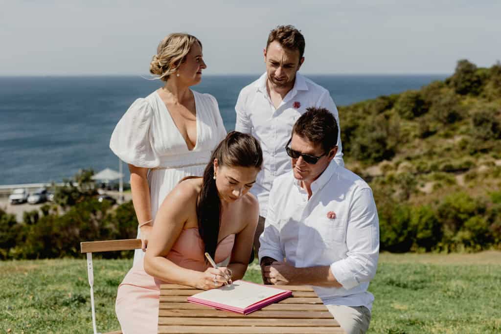 Witnesses signing paperwork while couple stand behind at  Newcastle coastal wedding